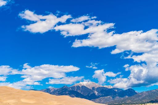 A stunning view of sand dunes under a vibrant blue sky with clouds, set against a backdrop of majestic mountains.