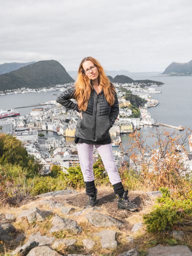 A Caucasian woman with long hair wearing glasses and posing in front of the city of Alesund, Norway