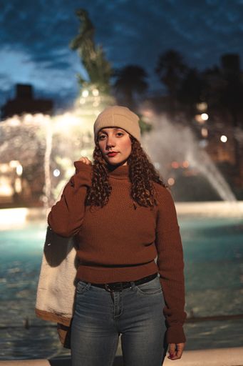 A vertical shot of a young female wearing a brown sweater and warm hat standing in front of a fountain