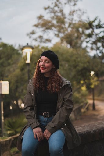 A vertical shot of a female wearing a khaki jacket and a black warm hat sitting in the park