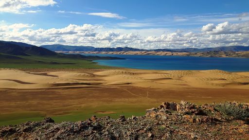 A panoramic shot of a sand landscape in front of the lake surrounded by hills under the cloudy sky