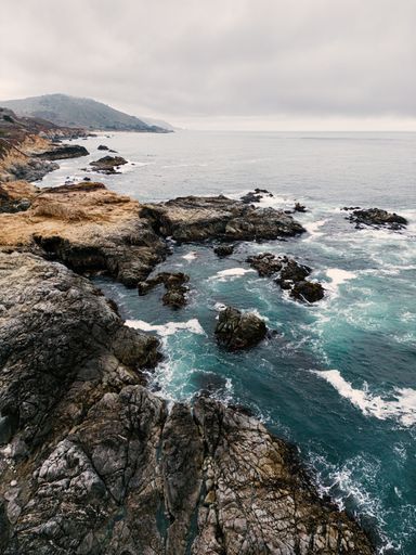 Majestic Big Sur coastline with waves crashing against rugged cliffs beneath a dramatic, cloud-filled sky.