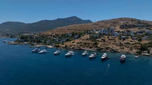 Aerial view of luxury yachts anchored in a serene bay near a coastal town with hills in the background under a clear blue sky.