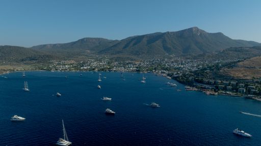 Aerial view of a serene bay with yachts and a coastal town under a clear blue sky, surrounded by lush mountains.