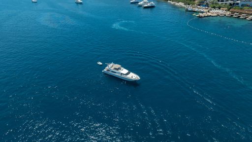 Aerial view of a yacht sailing on the clear blue waters near a coastal area with other boats and a resort visible in the background.