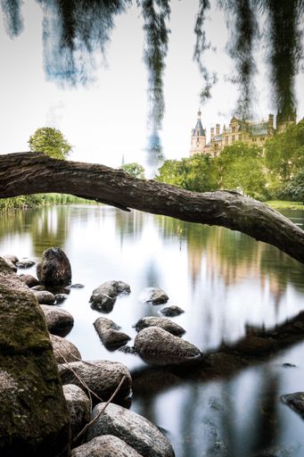 A vertical shot of a river with small stones and tree roots in it on a castle background