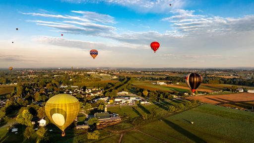 Aerial view of colorful hot air balloons floating over a scenic rural landscape under a clear blue sky.