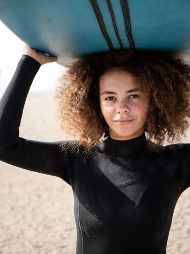 Young woman in wetsuit holding surfboard overhead on a sunny beach, smiling confidently.