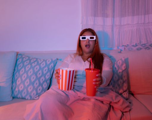 Young woman wearing 3D glasses, holding popcorn and drink, watching a movie at home, illuminated by colorful lights.