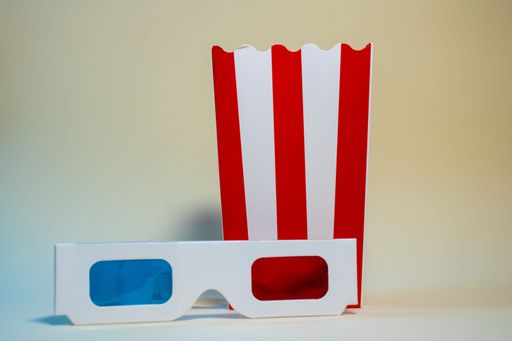 A pair of 3D glasses and a red and white striped popcorn box on a neutral background.