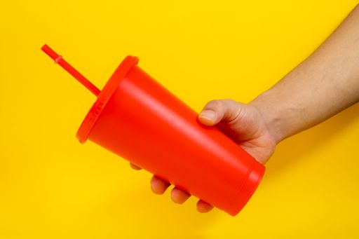 A hand holding a bright red cup with a straw against a vibrant yellow background.