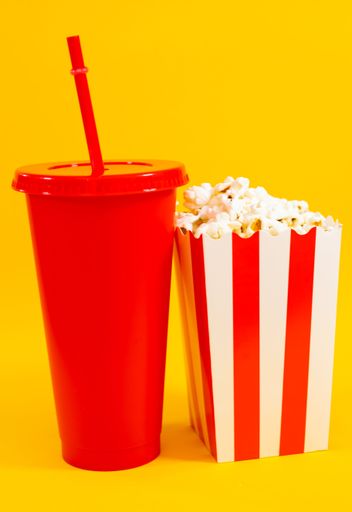 A red soda cup with a straw and a striped popcorn box on a vibrant yellow background.