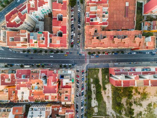 A top-down aerial view of a city intersection surrounded by red-roof buildings, streets, and parked cars, showcasing urban planning and architecture.