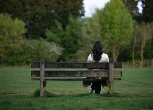 A woman with long dark hair sits on a wooden bench in a lush green park, surrounded by trees, enjoying a peaceful moment in nature.
