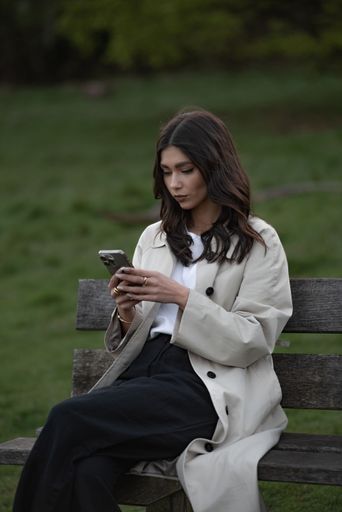 Young woman in a beige coat sitting on a park bench using a smartphone, surrounded by greenery, portraying modern lifestyle and technology.