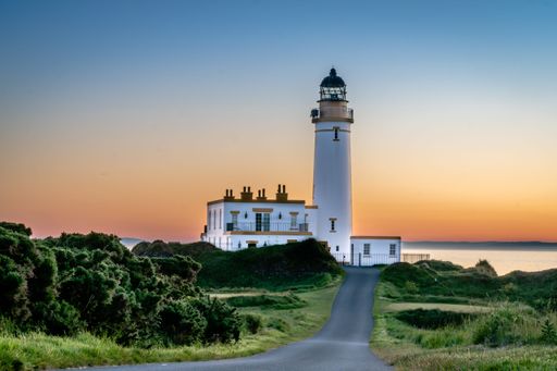 A scenic view of a lighthouse at sunset, with a clear sky and surrounding greenery, creating a serene coastal landscape.