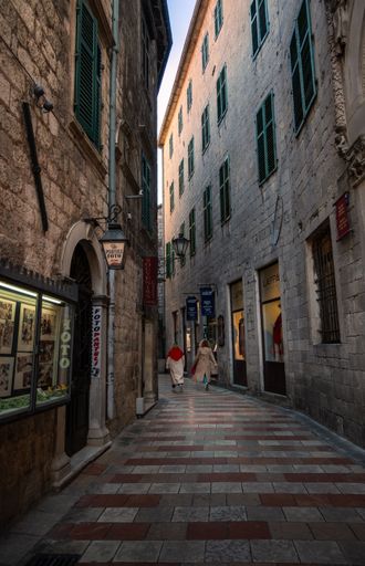 A narrow cobblestone street in Kotor, Montenegro with two women walking, surrounded by historic stone buildings and green shutters.