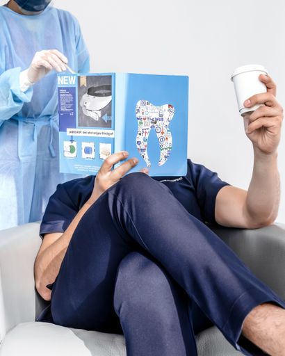 A dental professional in blue scrubs reads a magazine while holding a coffee cup, with a nurse in protective gear assisting.
