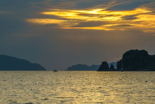 A serene sunset over a calm sea with silhouetted islands and a boat in the distance, creating a tranquil coastal scene.