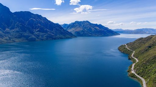 Aerial view of a scenic road winding along a stunning lake surrounded by majestic mountains under a clear blue sky.