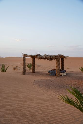 A serene desert scene with a rustic shelter and seating under a clear sky, highlighting the tranquil beauty of the sandy landscape.