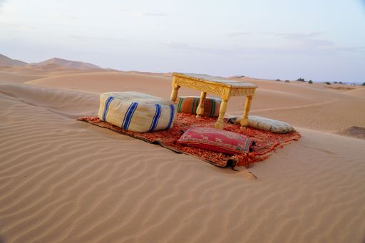 Traditional Moroccan seating arrangement with cushions and a table on sand dunes in the Sahara Desert under a clear sky.