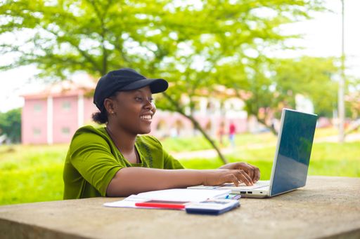 A pretty African female student happily doing her assignment with laptop, calculator and textbooks on a table in the campus park