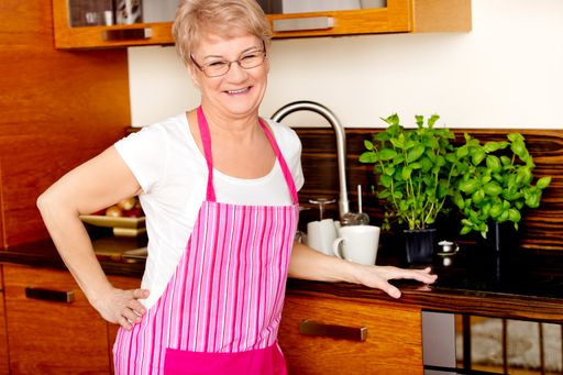 A cheerful elderly Polish female wearing an apron leaning against the counter in the kitchen