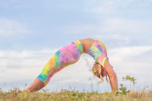 Young woman in bright tracksuit stands in a "bridge" pose on a green meadow, against the background of a blue summer sky. Outdoor gymnastics.