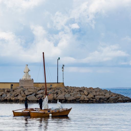 A small sailboat with two people navigating near a rocky breakwater under a cloudy sky in a tranquil harbor setting.
