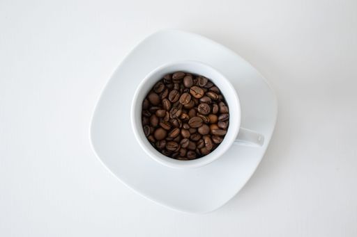 Top view of a white cup filled with coffee beans on a matching saucer against a clean white background