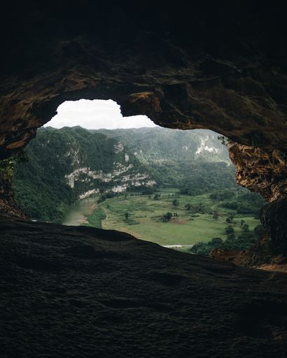 Scenic view of lush green valley through a cave opening with rugged rock formations and distant hills under a cloudy sky.
