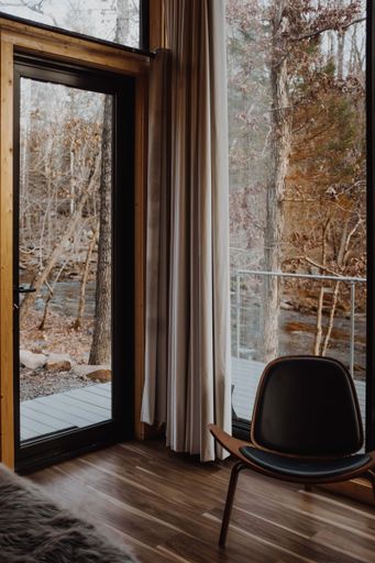 Cozy interior with a modern chair by a large window overlooking a serene wooded landscape and a flowing stream.