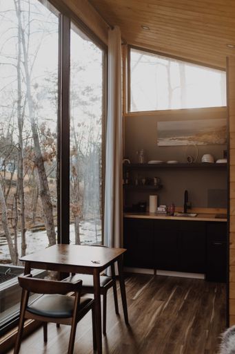 Cozy modern interior of a cabin with large windows overlooking a serene forest view, featuring wooden floors and minimalist decor.
