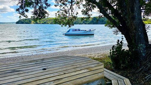 A serene lakeside view with a boat anchored near the shore, framed by trees and a wooden deck, under a partly cloudy sky.