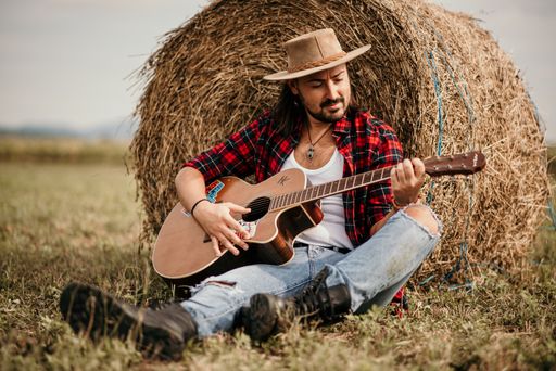 A young handsome Serbian male in a flannel shirt sitting leaned on a haystack and playing a guitar