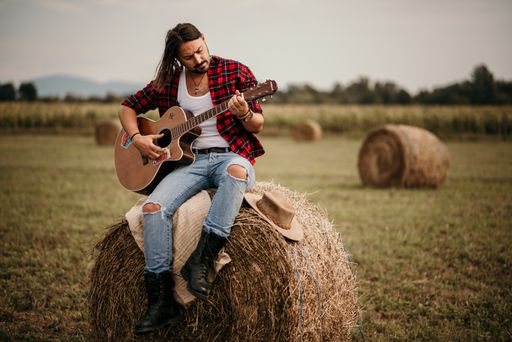 A young handsome Serbian male in a flannel shirt sitting on a haystack and playing a guitar
