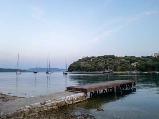 Scenic view of a tranquil bay with sailboats and a wooden pier under a clear sky, surrounded by lush greenery.