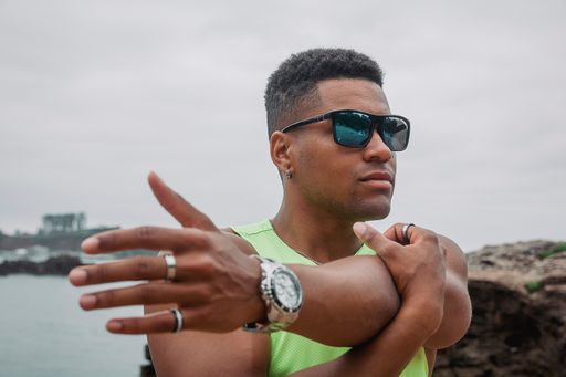 The young fit man with sunglasses and nose piercing stretching his arms near the ocean