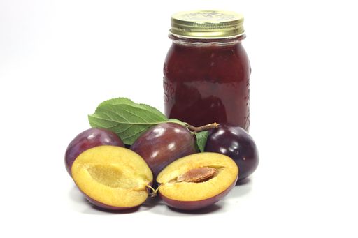 A glass container of black plum, surrounded by raw plums with a white background