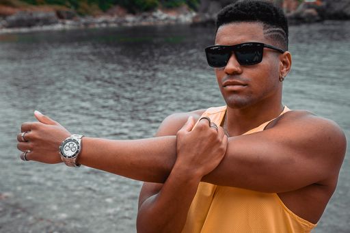The young fit man with sunglasses stretching his arms with the calm ocean in the background
