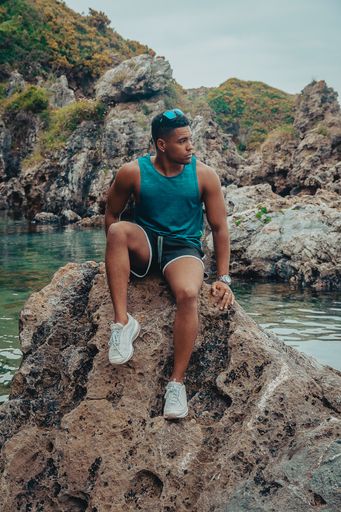 A young fit man with sunglasses sitting on the rock near the calm sea during daytime