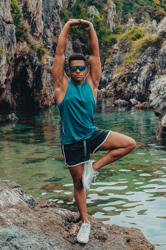 The young fit man with sunglasses doing yoga by balancing on one leg on the rock in the ocean