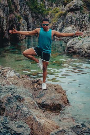 The young fit man with sunglasses doing yoga by balancing on one leg on the rock in the ocean
