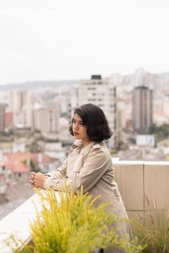 A vertical shot of the young woman in a cream coat looking from the balcony.