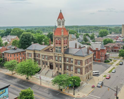 Aerial view of a historic courthouse with a clock tower in a small town setting, surrounded by trees and roads, captured on a cloudy day.