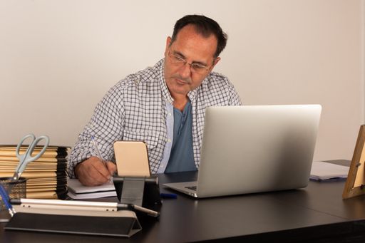 A middle-aged Caucasian man wearing glasses and taking notes sitting in front of the laptop