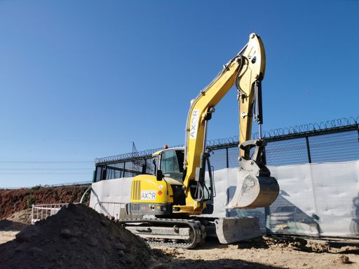 An excavator from the rental company AXOR parked next to a mound of dirt in a fenced work area