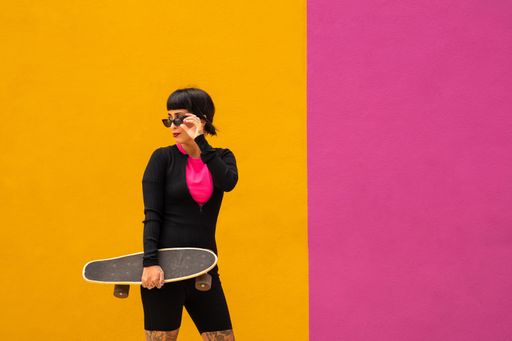 A beautiful young female in sunglasses posing with a skateboard on pink and yellow background