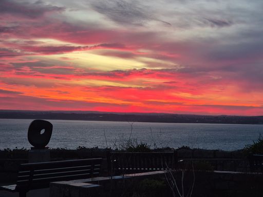 Vibrant sunset over the ocean with silhouetted sculpture and benches in the foreground, creating a serene coastal scene.